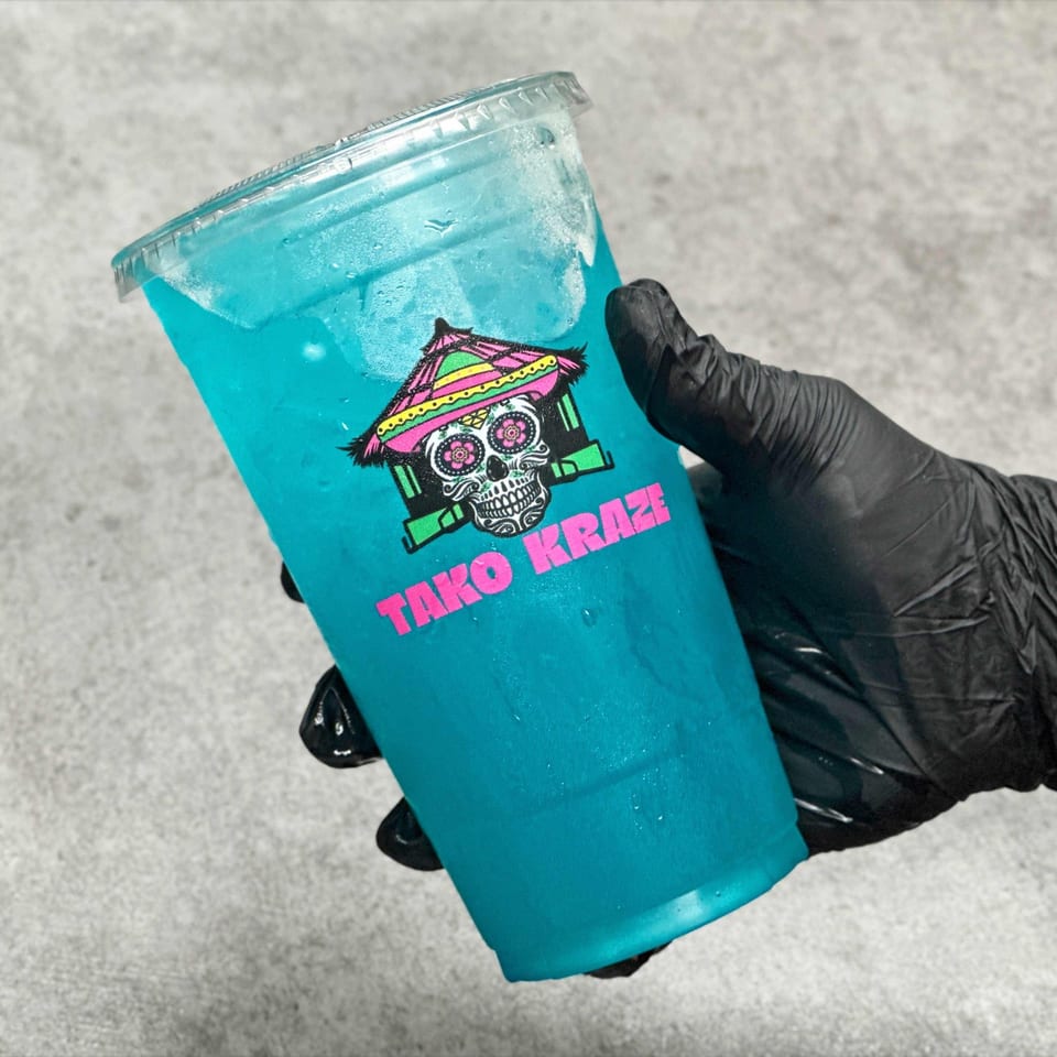 Hand in black glove holding a clear cup of bright blue drink with the Tako Kraze logo.