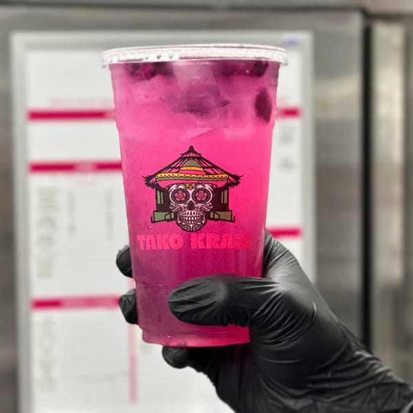 Hand in black glove holding a vibrant pink Tako Kraze drink with ice and fruit pieces.