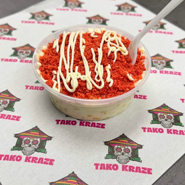 Tako Kraze elote in a clear cup, topped with red chili powder and white crema, with a spoon.